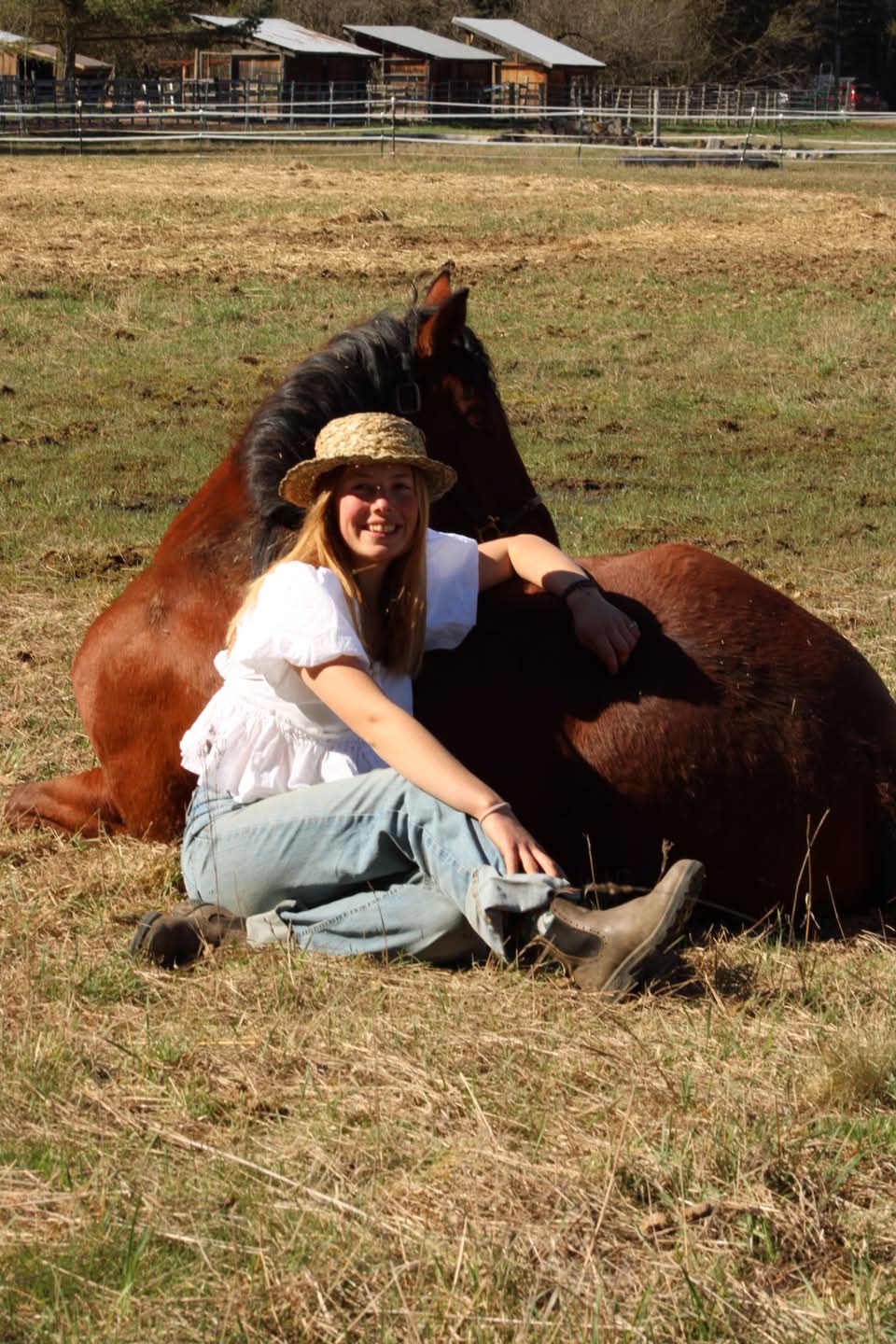 Maryah ’28 in a straw hat sitting in the grass beside Delilah as the mare lies down