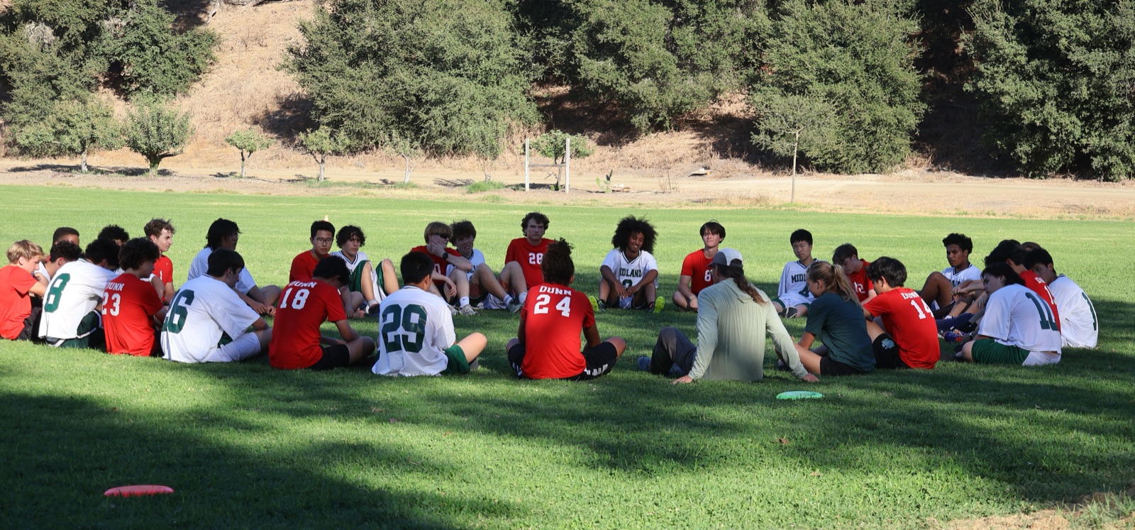 Midland and Dunn Ultimate Frisbee players sitting together in a circle on a grass field