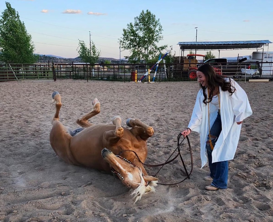 Peaches rolling in the sand of an arena while Bri ’29 laughs and holds the reins